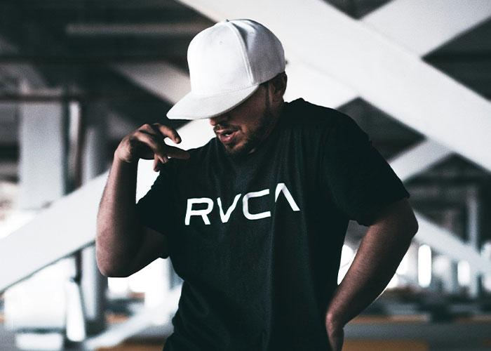 Man in RVCA shirt and white cap performing a dance move in an industrial setting.