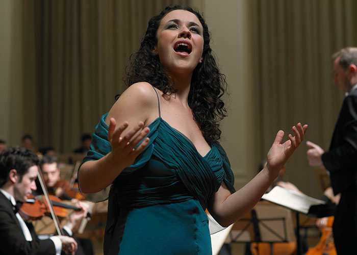 A woman singing passionately on stage with an orchestra, representing brightest kids' potential and success.