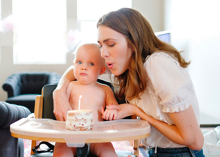 A mother and baby at a birthday celebration, with the mother blowing out a candle on a small cake.