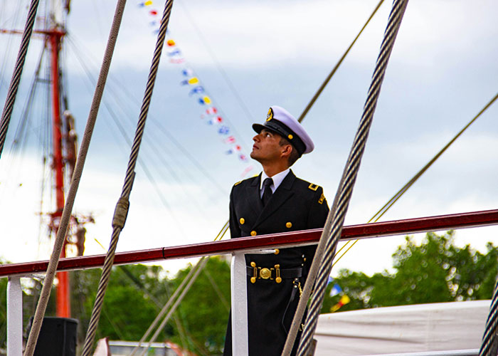 A naval officer in uniform stands on a ship deck, surrounded by ropes and flags, looking upwards on a cloudy day.