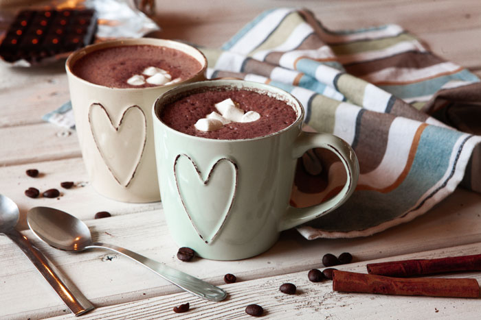 Two heart-adorned mugs of hot chocolate on a wooden table with scattered coffee beans and cinnamon sticks.