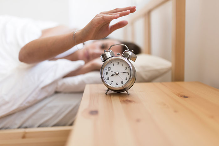 A woman reaching to turn off alarm in bed, illustrating morning wake refusal.