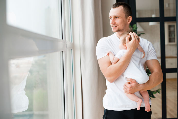 Man holding a baby, standing by a window, contemplating morning wakeup routines, while gazing outside.
