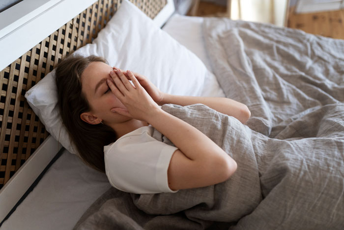 Woman in bed with covers pulled up, hands on face, subtly refusing to wake up in the morning.