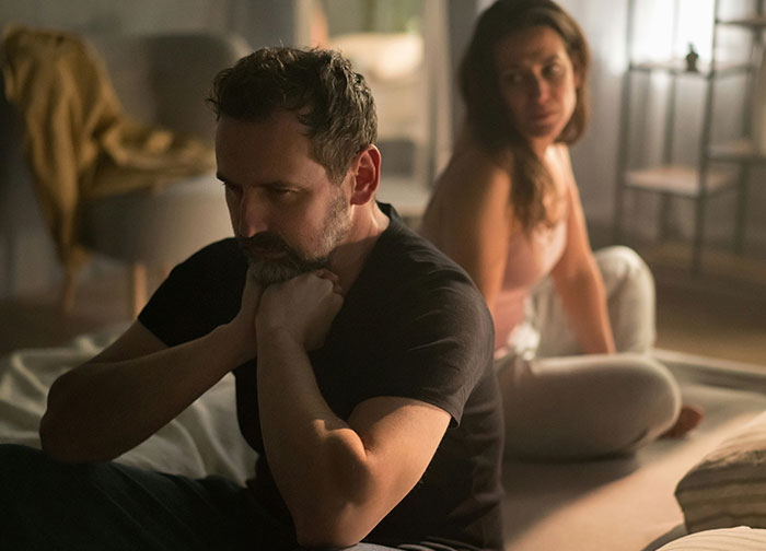 A concerned husband sits with his wife, questioning her explanation of an apparent hickey.