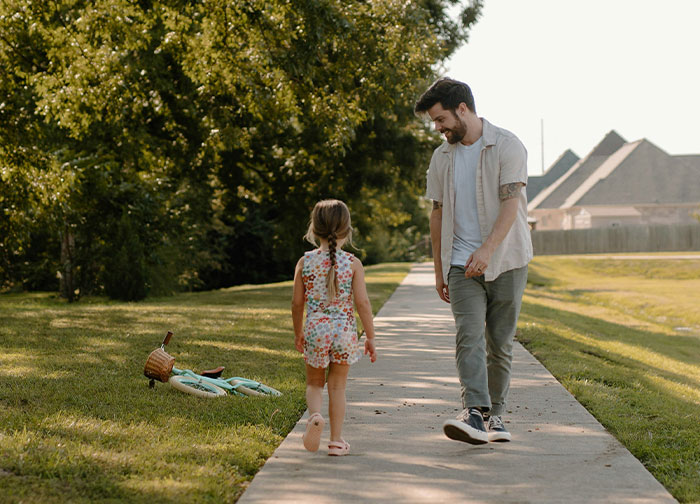 Dad and daughter walking on a sunny path, bike lying on grass, surrounded by trees.