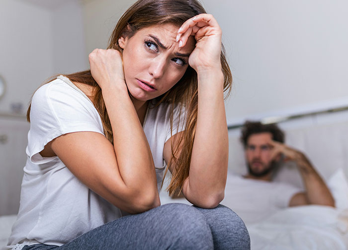 Woman looking concerned on bed, husband in background, related to hickey and bug bite suspicion.