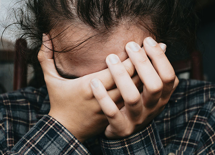 A woman covering her face with her hands, wearing a plaid shirt, possibly stressed over a situation involving a hickey.