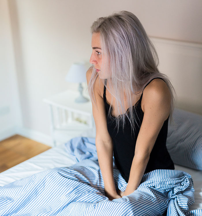 A woman with silver hair in a bedroom setting, sitting on a bed, looking away.