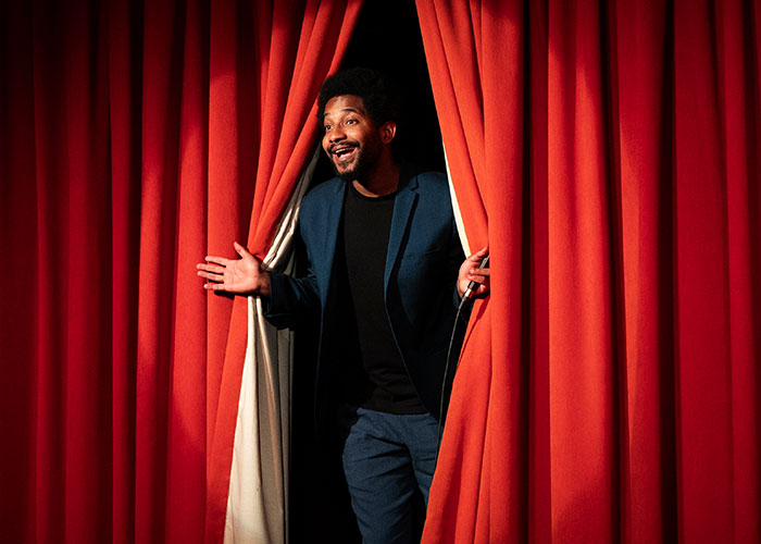 Person in a blue suit peeking from red theater curtains during a wild audition moment.