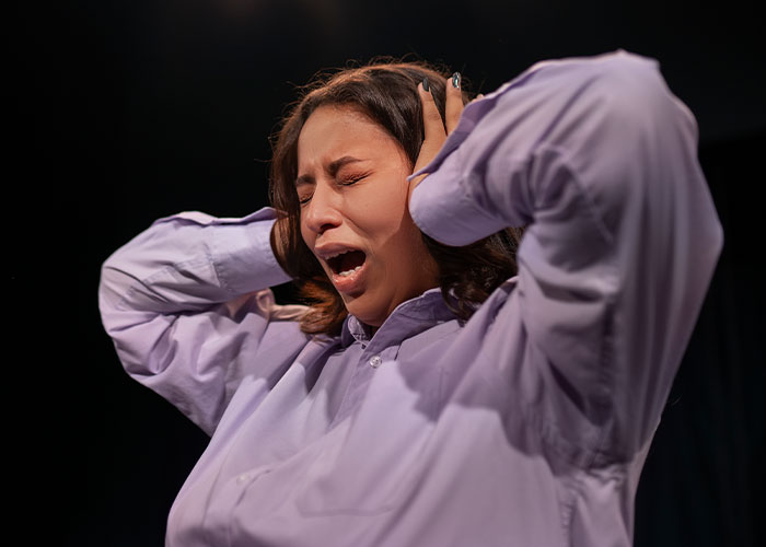 Person in emotional performance during a wild audition, wearing a purple shirt with hands over their ears.