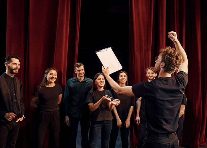 Actors rehearsing on stage, holding a clipboard in an audition setting with red curtains in the background.