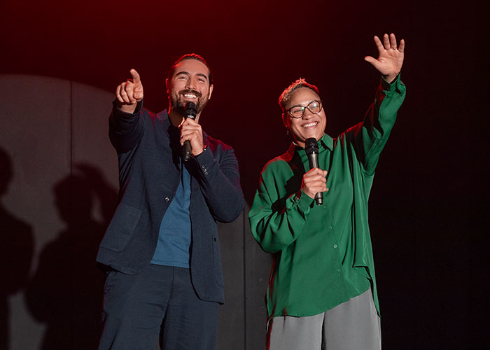 Two people on stage during an audition, smiling and holding microphones under stage lights.