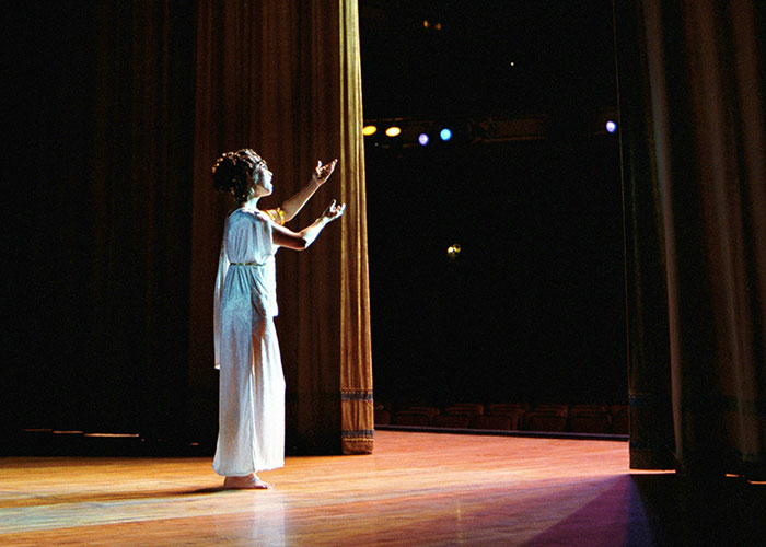 Performer on stage during an audition, dressed in a white gown with dramatic lighting highlighting the scene.