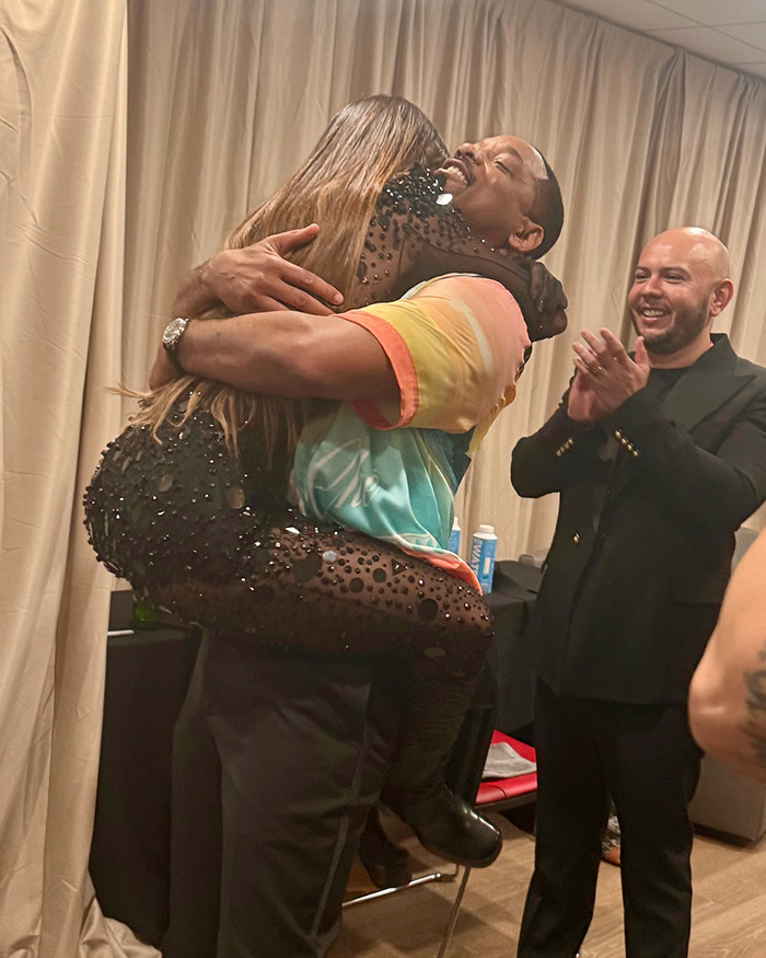 Man hugging a woman in a colorful shirt, with another man clapping, related to Will Smith controversy. Man hugging a woman in a colorful shirt, with another man clapping, related to Will Smith controversy.