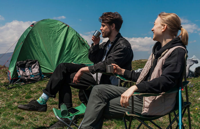 Two people sitting outside a green tent enjoying drinks in a campsite under a blue sky.