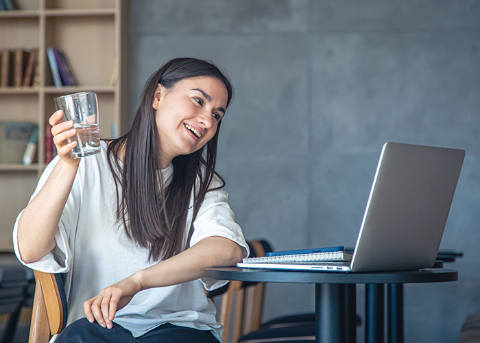 College student pregaming midterm online, holding a drink, and smiling at her laptop.