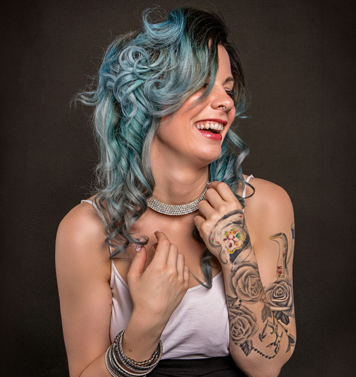 Woman with blue hair and tattoos laughing, wearing a white top and necklace.