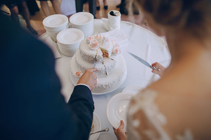 A wedding cake being cut by a couple at a wedding reception with stacks of plates nearby.