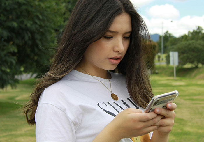 Young woman in a white shirt looking at her phone, standing in a park.