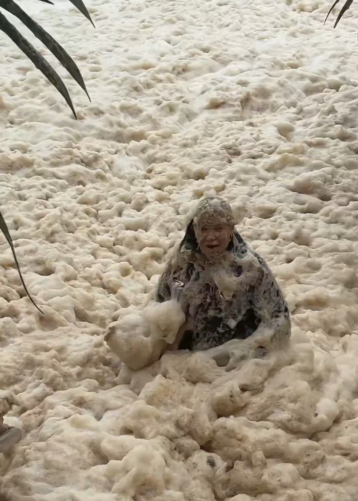 Woman performs stunt in sewage-filled sea foam, partially covered, amidst palm leaves, sparking outrage online. Woman performs stunt in sewage-filled sea foam, partially covered, amidst palm leaves, sparking outrage online.