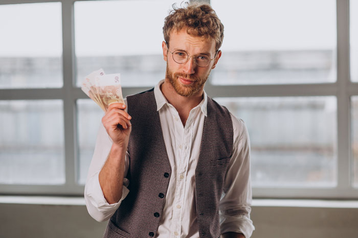 Man holding cash, wearing glasses and vest, in a bright room. Keywords: sibling, demands, 20K, surgery.