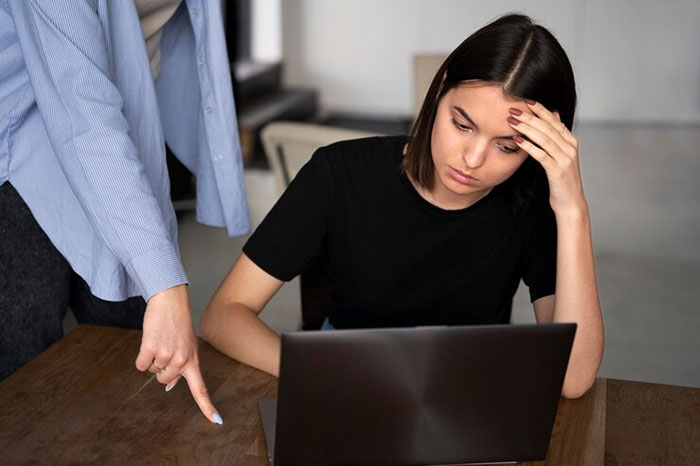Woman looking embarrassed at laptop while colleague points at screen in office setting.