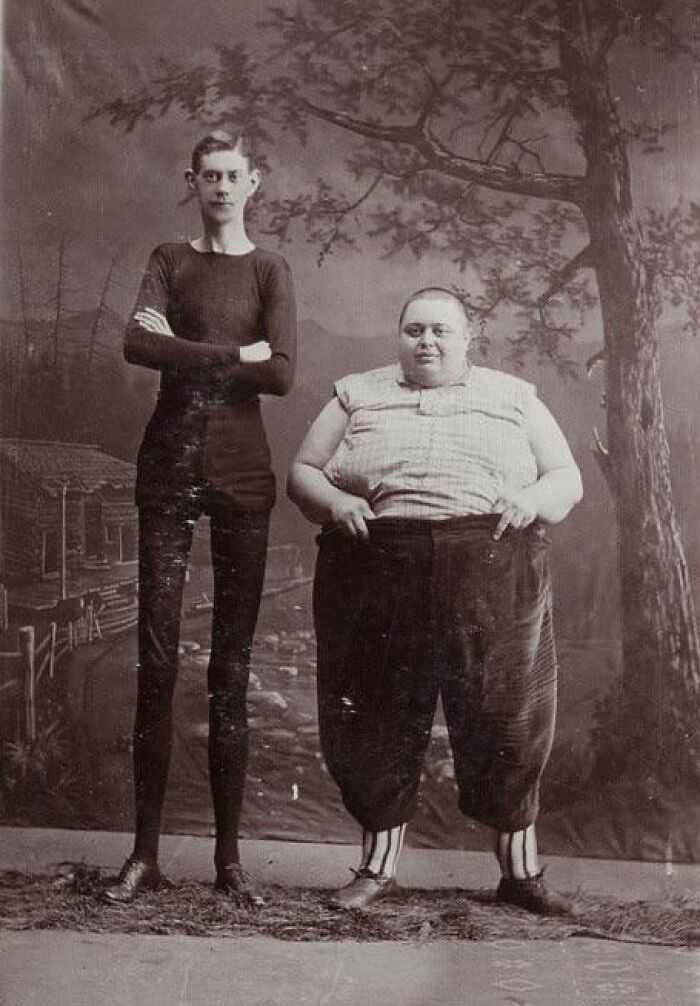 Two men from the past posing together, one tall and thin, the other short and stout, in an unsettling vintage scene.