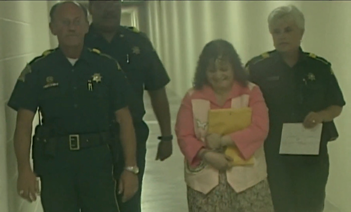 Woman escorted by officers in a hallway after parole denial for pop star assassination.