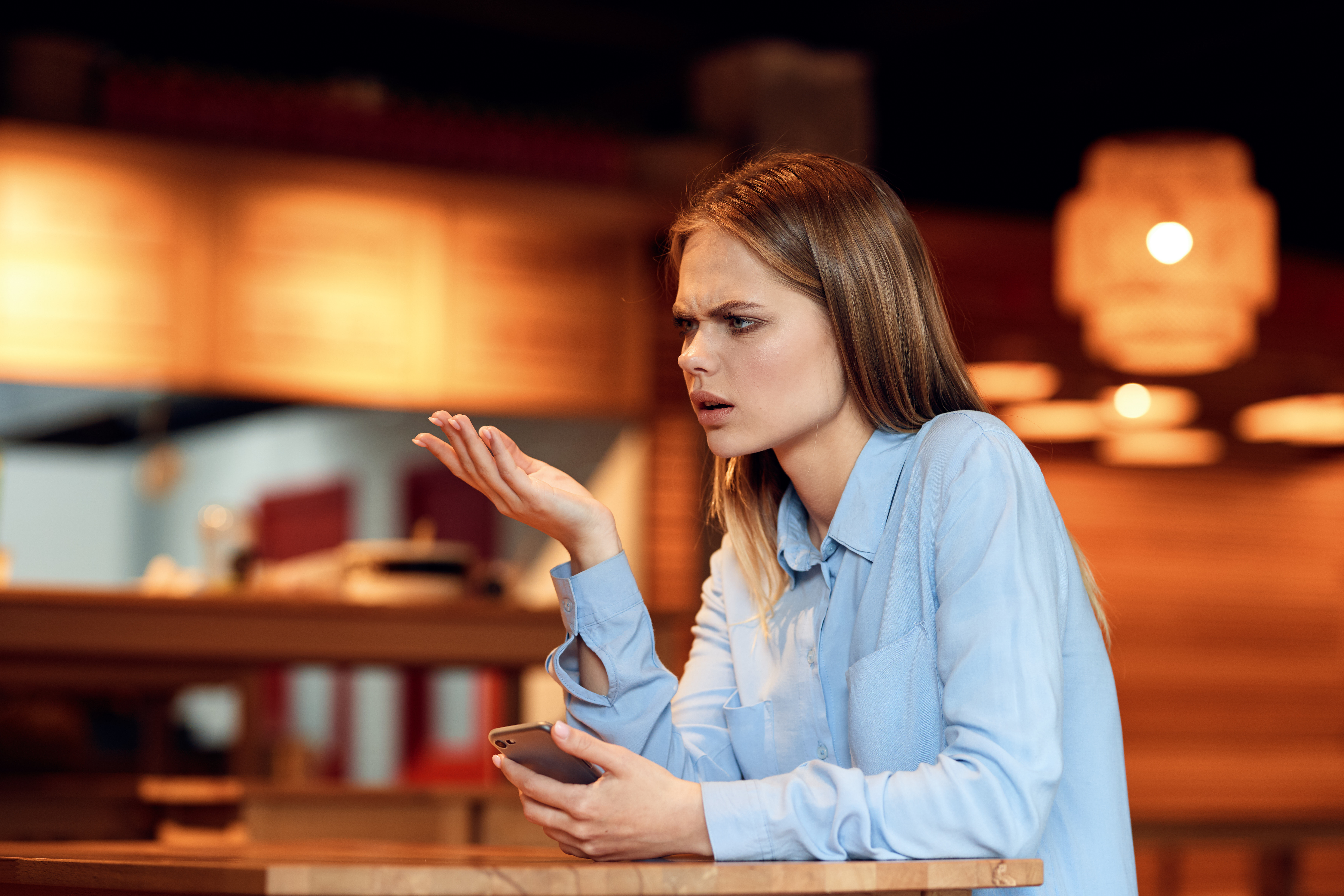 Confused woman in a blue shirt holding a phone at a table, under warm lighting.