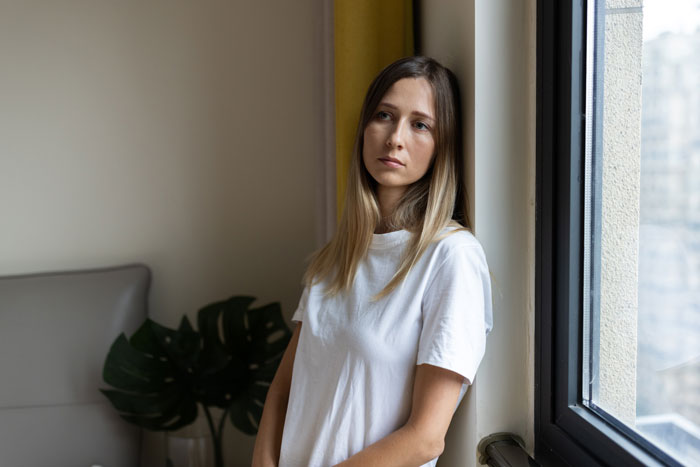 Woman in a white shirt leaning by a window, looking thoughtful.