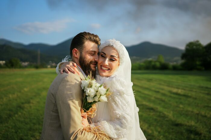Bride and groom embrace in a scenic field, smiling after a wedding ceremony, with mountains in the background.