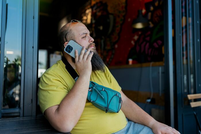Man in yellow shirt, sitting outdoors with phone, expressing frustration.