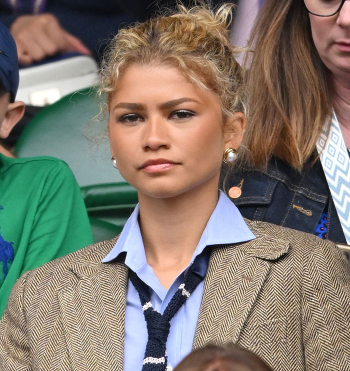 Woman in a herringbone blazer and blue shirt at an outdoor event, looking serious.