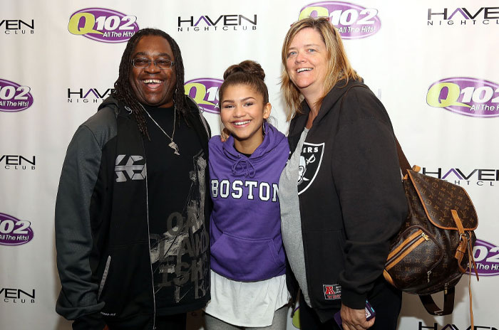 Zendaya with her family at a Q102 event, wearing a purple Boston hoodie, smiling between her parents.