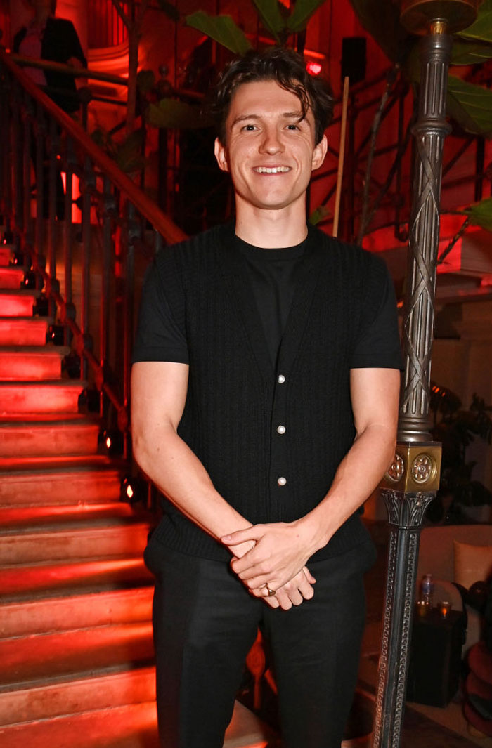 Tom Holland standing in a dimly lit setting, leaning against a staircase, related to Zendaya's family context.