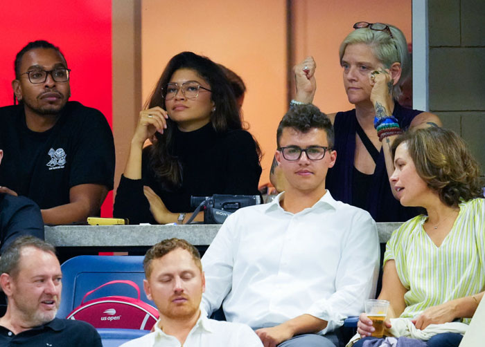 Zendaya seated with family at an event, wearing glasses and a black outfit, engaging with those around her.
