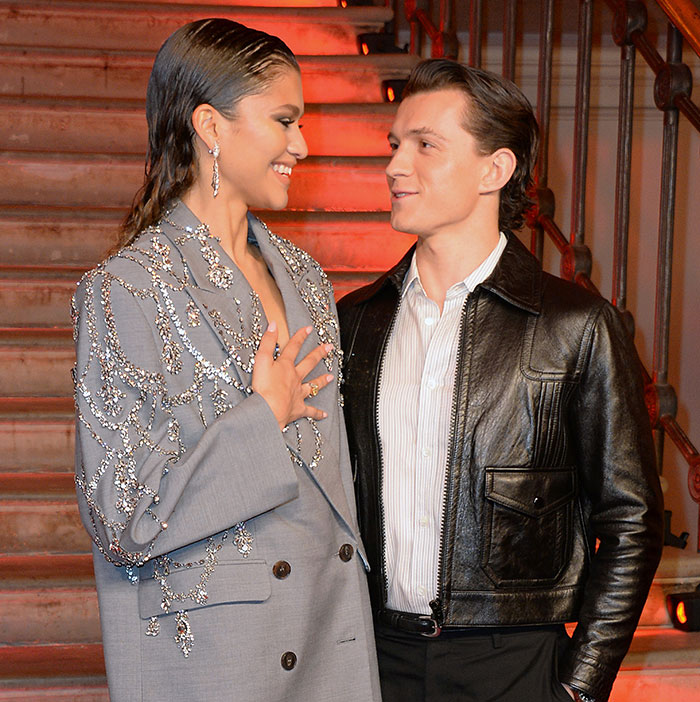 Zendaya and Tom Holland smiling at each other, dressed in formal attire, standing near stairs.