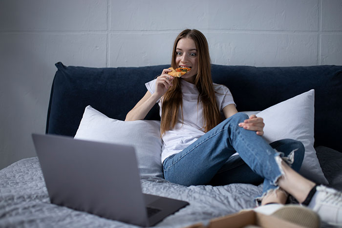 Young woman eating pizza on bed while watching a detailed breakdown about dating perpetually single women on laptop.