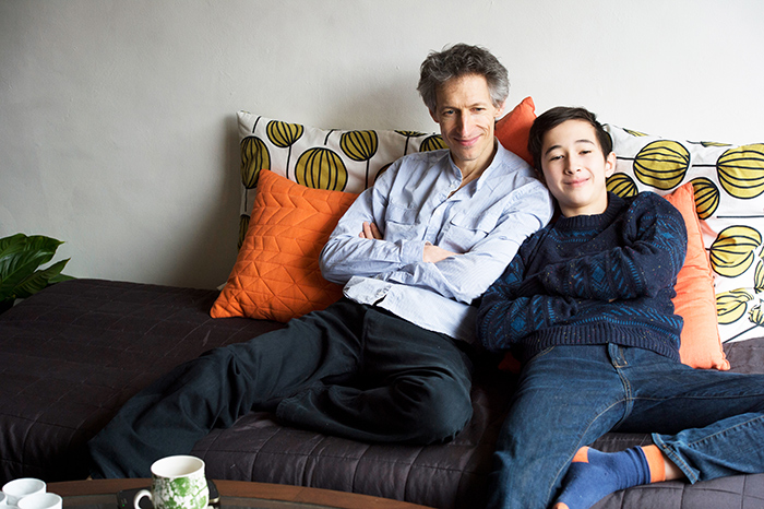 Father and teen boy lounging on a couch, snuggling against colorful cushions in a cozy living room.
