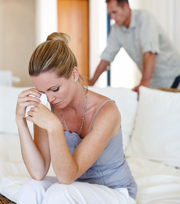 Woman sitting on couch tearfully holding tissue, feeling guilty about late father’s estate while man stands in background. Woman sitting on couch tearfully holding tissue, feeling guilty about late father’s estate while man stands in background.