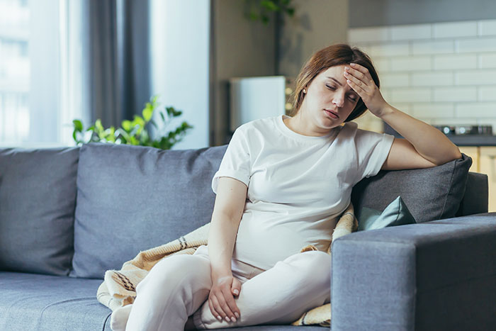 Pregnant woman on a couch, looking stressed in a modern living room setting.
