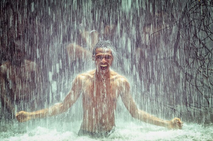 Man standing under heavy waterfall, appearing enthusiastic, showcasing a thrilling movie moment that's terrible in real life.
