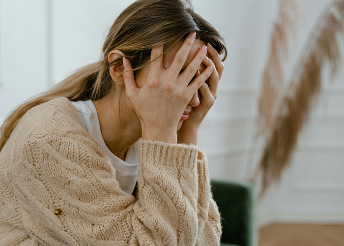 Woman in a beige sweater sitting with face in hands, appearing stressed, in a bright room with decorative plants.