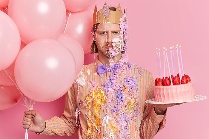 Man standing with balloons, covered in cake and holding a birthday cake, wearing a crown against a pink background.