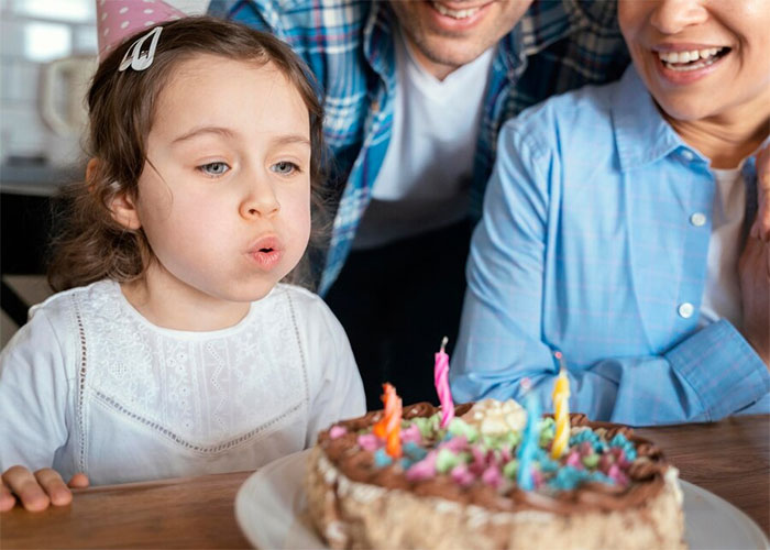 Child blowing out candles on birthday cake, surrounded by smiling adults.