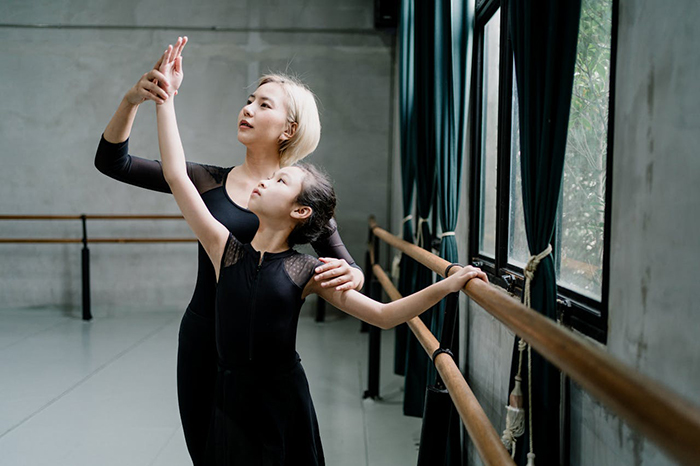 Dance teacher guides young girl at the barre in a ballet studio.