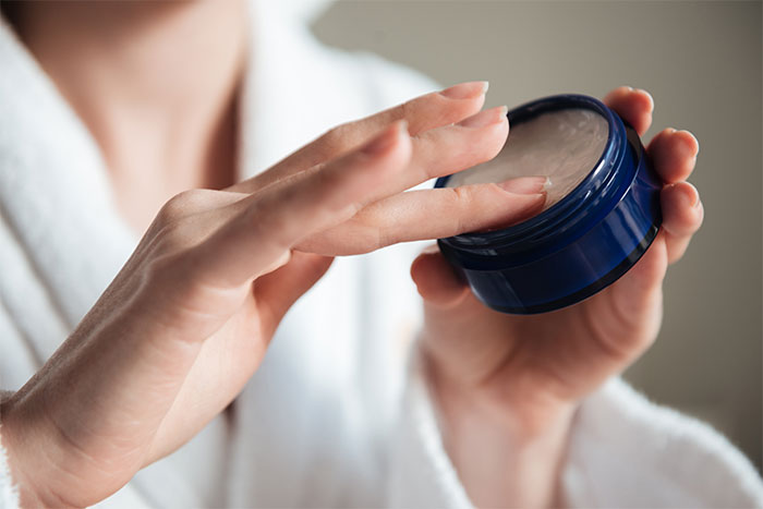 Hands applying cream from a blue container, focus on skin care routine.
