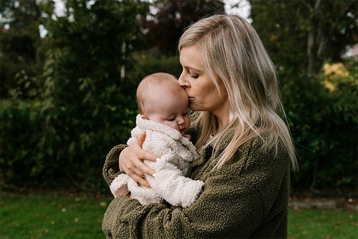 Mother kissing baby on forehead in a park setting, expressing warmth and affection.