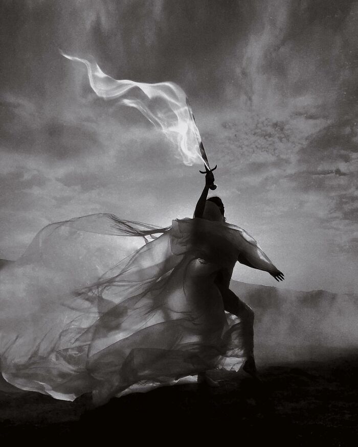 Dark escapism photography with a figure wielding a flaming sword in a flowing, ethereal outfit against a cloudy sky.